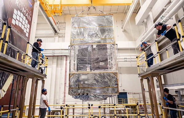 A large, rectangular satellite component being carefully lifted by a yellow overhead crane inside a spacious industrial facility. Six technicians wearing helmets observe and guide the process from elevated platforms, with a banner labeled 