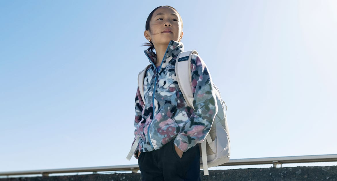 A little girl standing against a blue sky wearing a fleece jacket and a backpack.