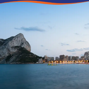 Coastal view featuring a large rock formation, city lights, and a calm sea at dusk.