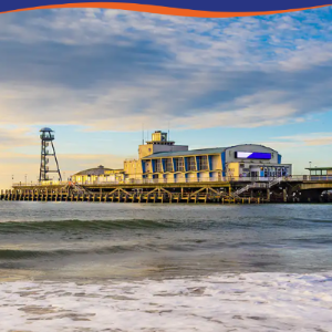 A seaside pier with a building and a watchtower under a cloudy sky at sunset.