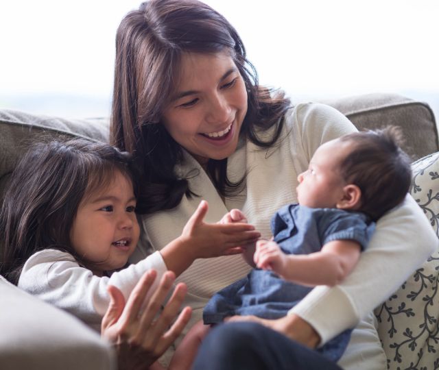 Mom sitting on the couch holding her infant and toddler.