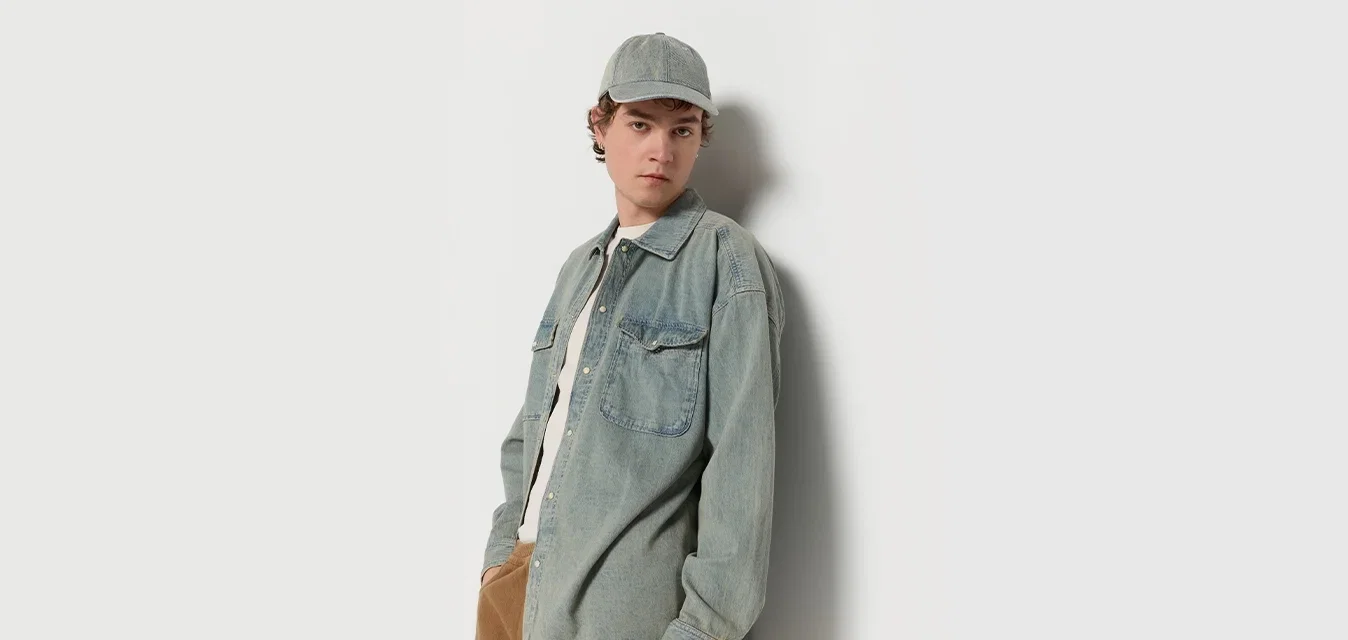 Lichtblauwe denimshirt met twee borstzakken, gecombineerd met een beige pet en bruine broek. Het shirt heeft een losse pasvorm en knoopsluiting.