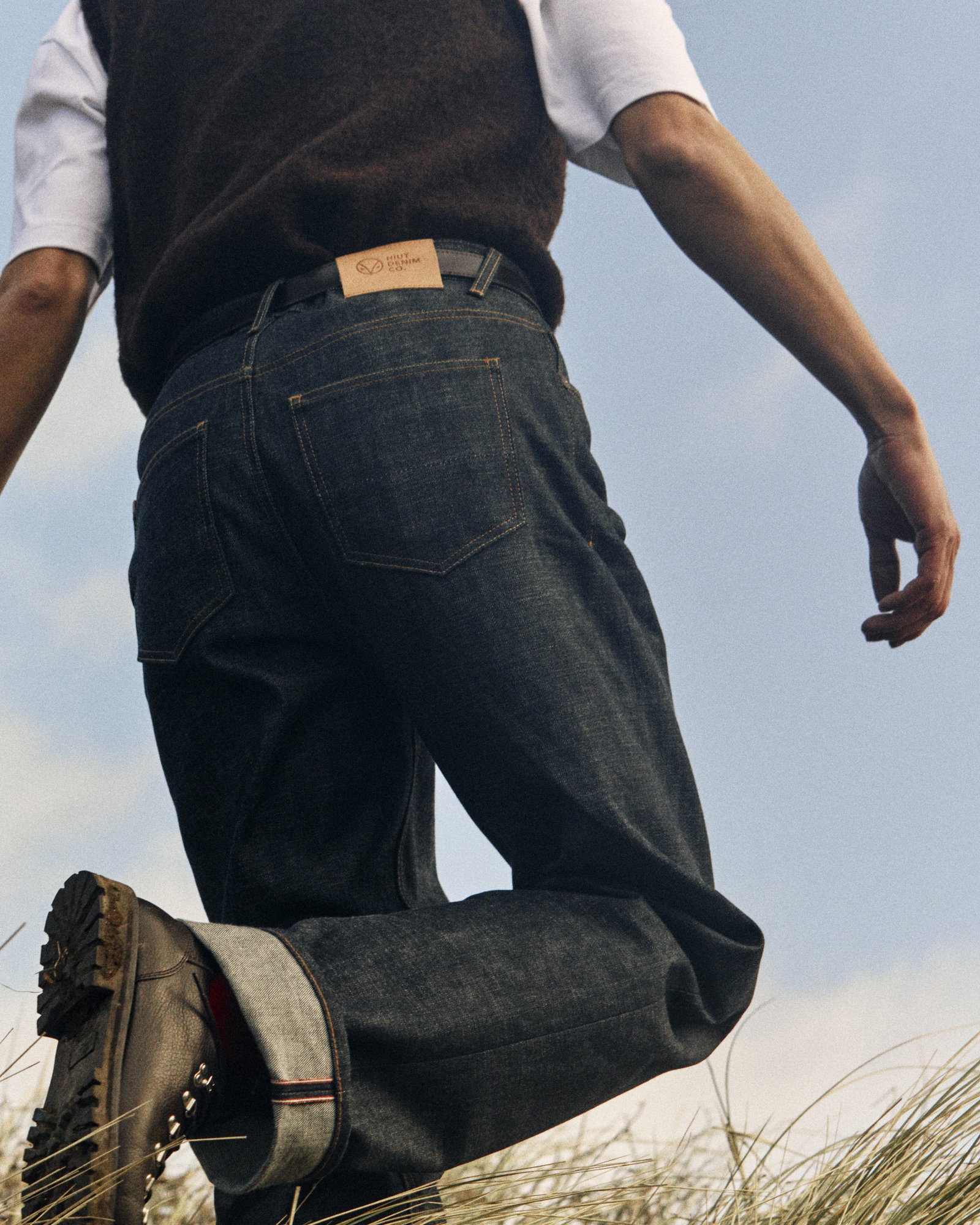 From behind image of man walking through dunes. He wears Miles selvedge denim Hiuts, white t-shirt, and wooden brown vest.