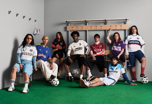 Group of models wearing retro-inspired MLS archive jerseys in a locker-room setting, seated and standing around benches with soccer balls on green turf. Jerseys represent multiple clubs.