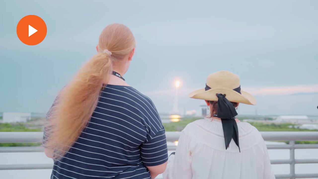 Two women stand on an observation deck watching a distant rocket launch from a railing, with a video play icon above