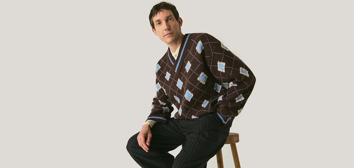 Man zittend op een houten kruk, gekleed in een bruine trui met blauwe ruitpatronen en zwarte pantalon met krijtstreep, tegen een effen achtergrond.