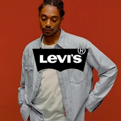Man met ingevlochten haar, gekleed in een licht denim overhemd en een ketting, staand met één hand op zijn heup tegen een effen oranje achtergrond.