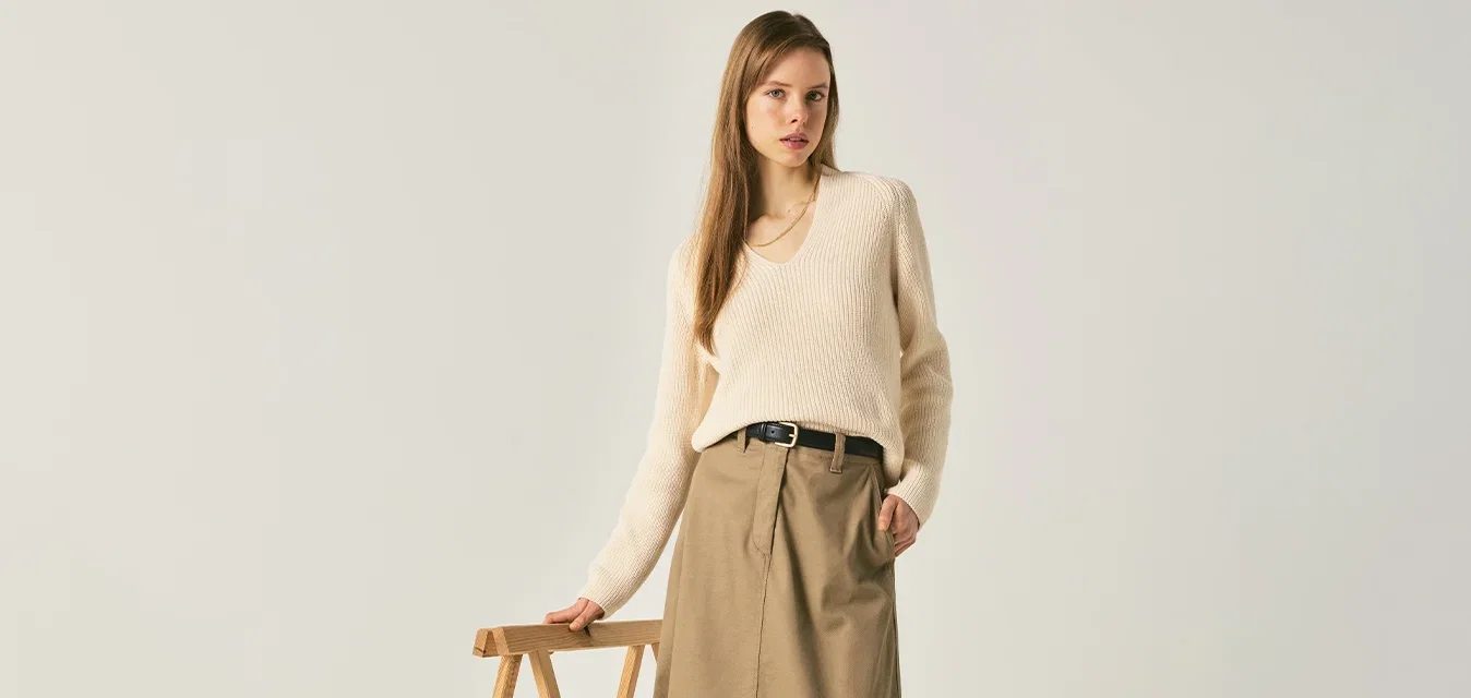 Frau trägt cremefarbenen Pullover, beigen Midirock und schwarze Loafer, steht drinnen und hält mit einer Hand ein hölzernes Sägepferd.