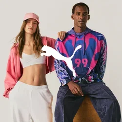Eine Frau in weißen Hosen, Crop-Top und pinker Jacke steht neben einem Mann, der auf einem Hocker sitzt und einen bunten Hoodie und dunkle Hosen trägt.