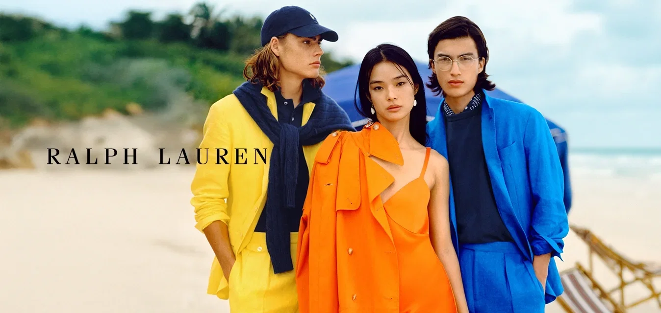 Drei Models stehen am Strand. Eine trägt einen gelben Anzug mit einem Marineblauen Pullover, die zweite ein oranges Kleid mit passender Jacke und die dritte einen blauen Anzug.
