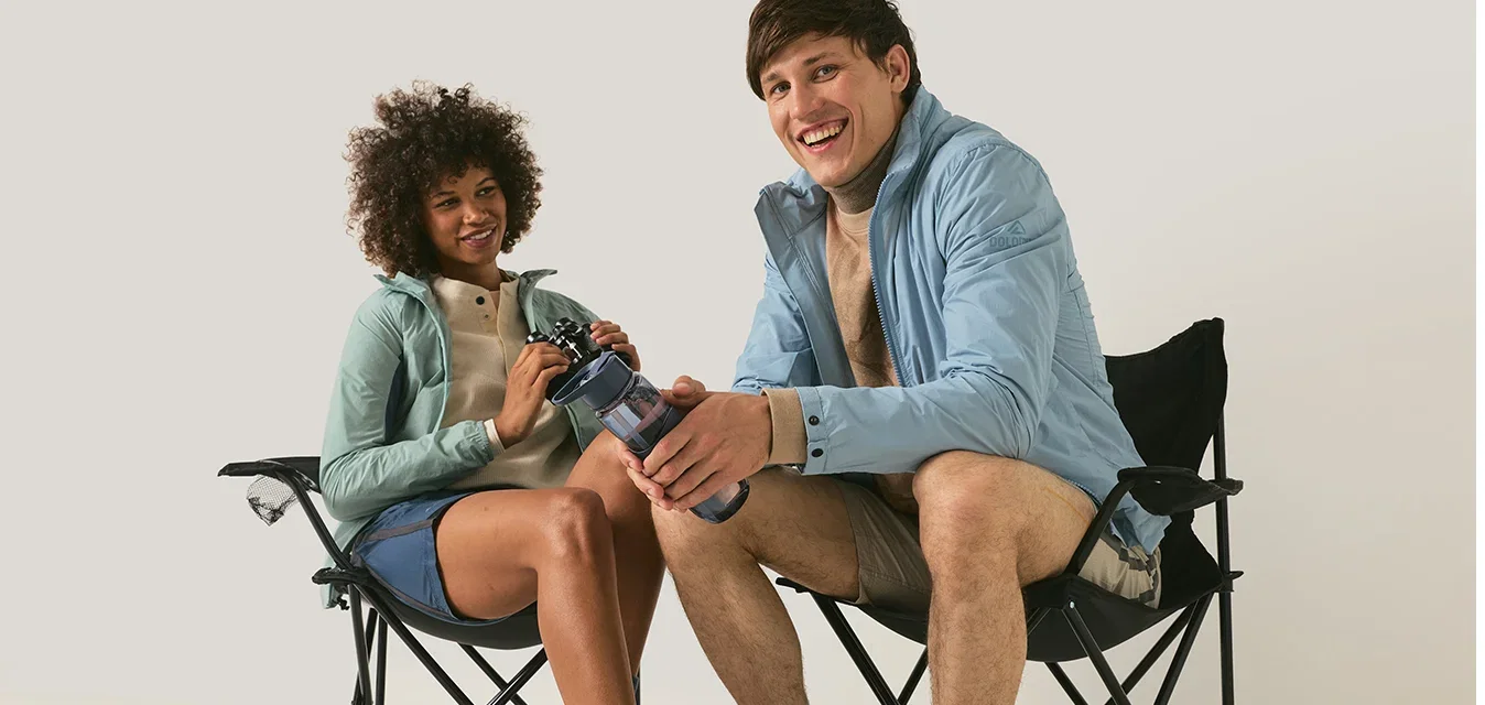 Zwei Personen in Outdoor-Kleidung sitzen auf Klappstühlen, halten und reichen eine Wasserflasche weiter, lächeln und schauen in die Kamera.