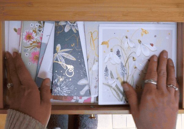Hands opening a wooden drawer filled with beautifully illustrated Whistlefish greeting cards featuring floral designs, gold foil details and seasonal artwork.