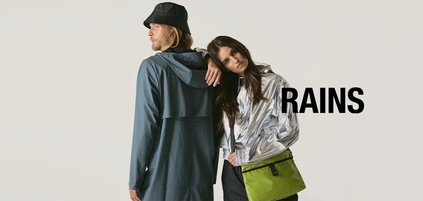 Een man in een blauwe regenjas en een zwarte bucket hat staat rug aan rug met een vrouw in een zilveren jack, terwijl ze haar hoofd op zijn schouder leunt en een groene tas draagt.