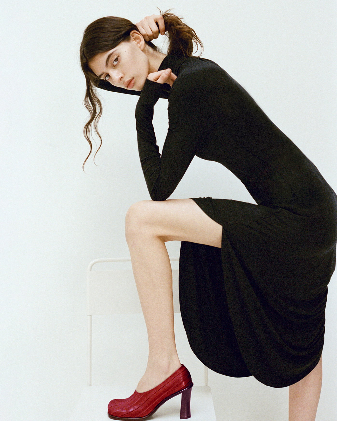 Image of model in a bright white room lifting her hair and placing leg on a chair while wearing the Imogen Dress in Soft Jersey in Black and the Esme 90MM Pumps in Eel in Red
