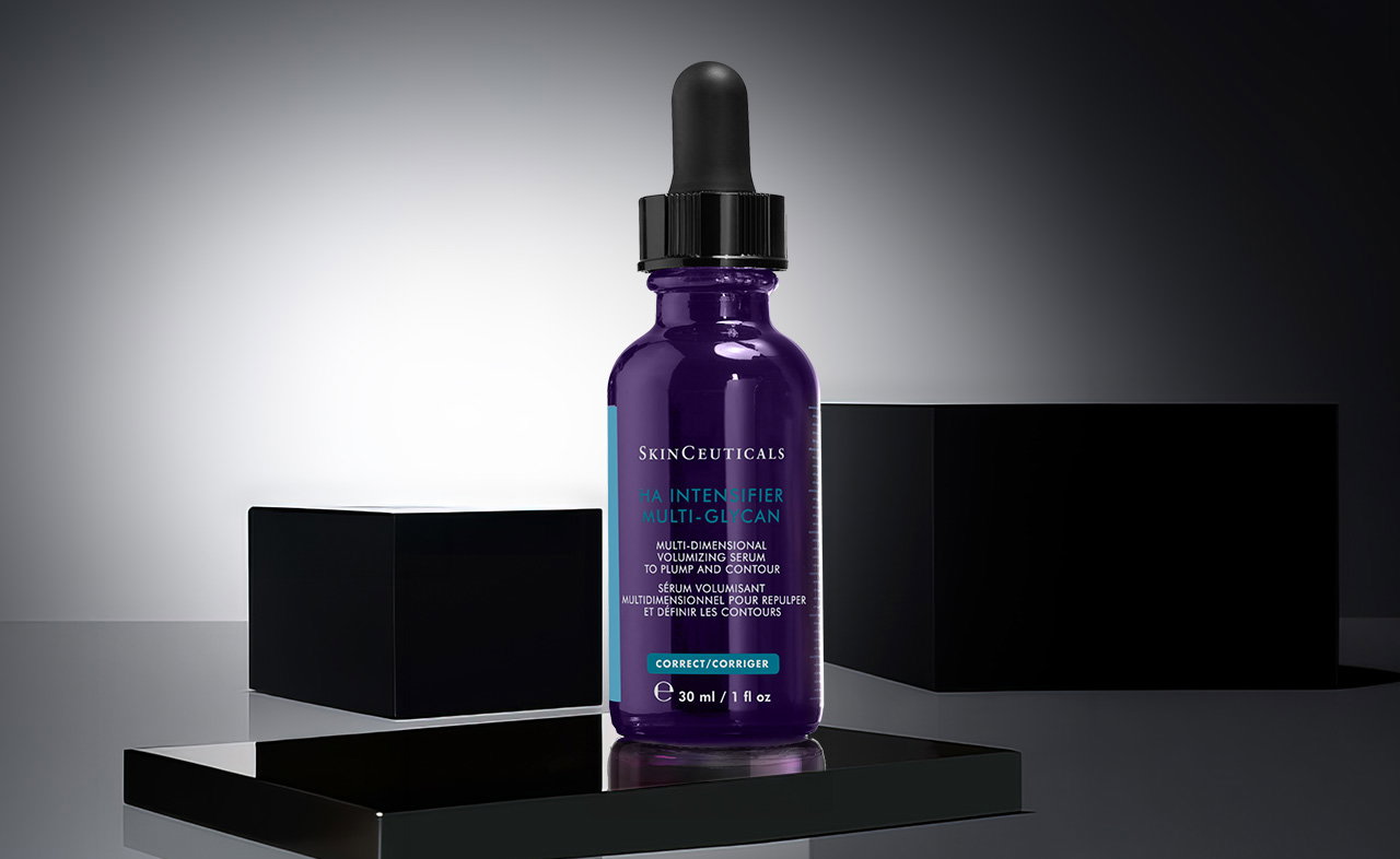 Eine einzelne SkinCeuticals HA Intensifier Multi-Glycan Serum-Flasche mit violettem Glas und schwarzem Pipettenverschluss auf einem schwarzen Podest mit zwei schwarzen Würfeln im Hintergrund.