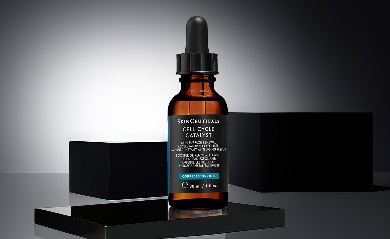 Eine einzelne SkinCeuticals Cell Cycle Catalyst Serum-Flasche mit braunem Glas und schwarzem Pipettenverschluss auf einem schwarzen Podest mit zwei schwarzen Würfeln im Hintergrund.