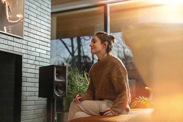 Young woman listening to a black Klipsch Sevens II speaker in a brightly-lit living room.