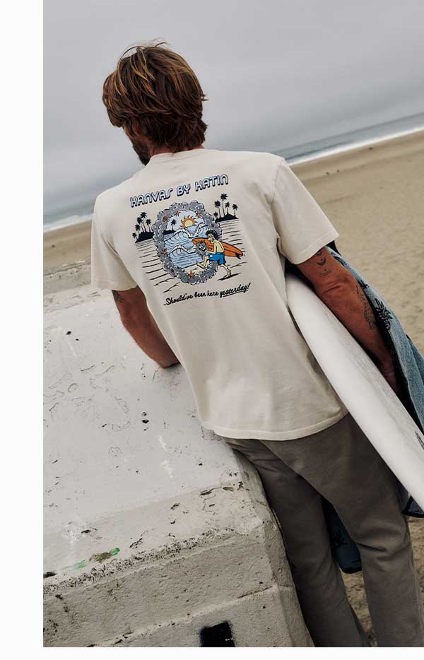 Back view of a man wearing a cream Kanvas By Katin graphic t-shirt, holding a surfboard at the beach