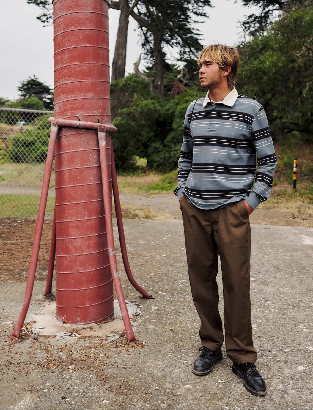 Man wearing a Katin striped rugby polo and brown pants standing beside a red water tank outdoors
