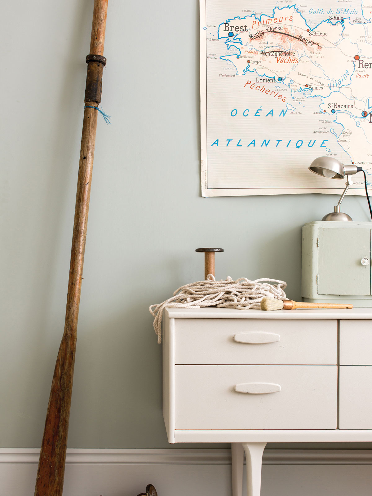 Vintage nautical-themed corner with a wooden oar, rope, map and white cabinet.