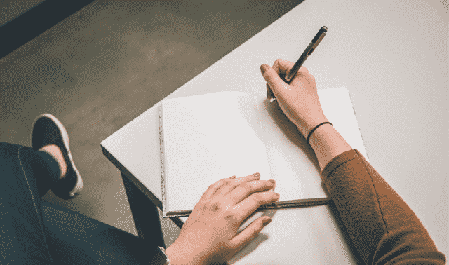 Person seated at a table, writing in an open notebook with a pen.