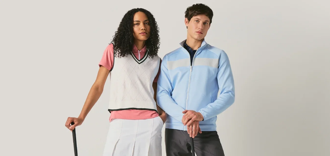 Twee golfers, een vrouw in een witte rok en vest, een man in een blauwe jas en zwarte broek, staande met golfclubs tegen een effen achtergrond.