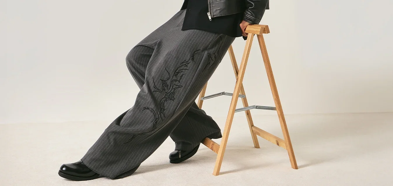 Persoon die een zwart leren jasje, brede grijze broek, zwarte schoenen en een zonnebril draagt, leunend op een houten vouwkruk tegen een effen achtergrond.