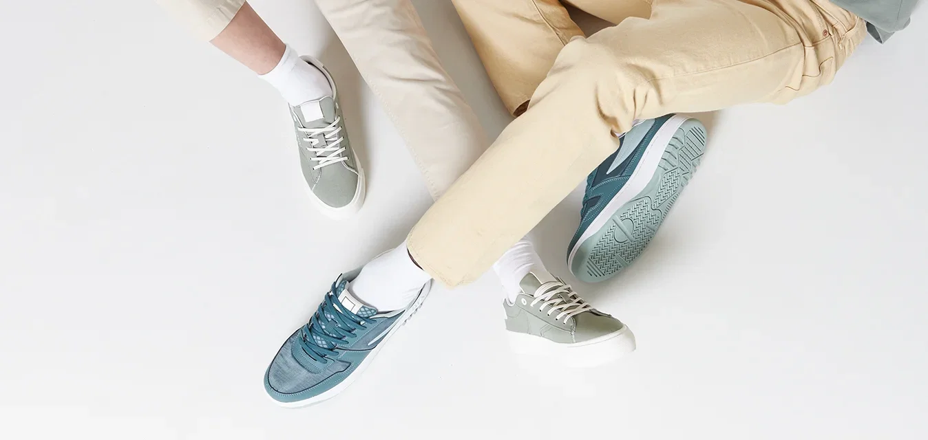 Twee personen zitten op een witte ondergrond, gekleed in een lichtgroen topje en beige broeken. Het schoeisel bestaat uit gestructureerde sneakers in twee kleuren, teal en grijs.