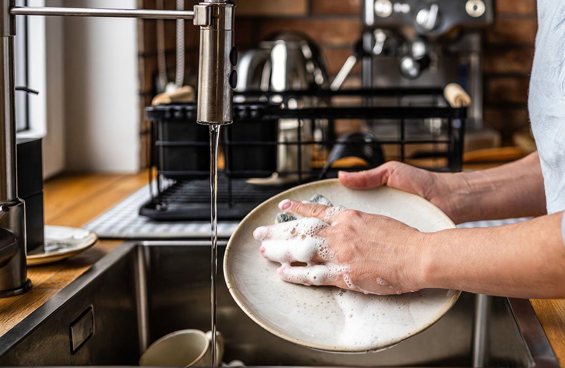 Person pre rinses a plate under running water beside a dish rack