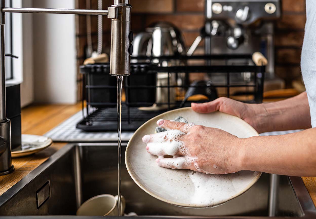 Person pre rinses a plate under running water beside a dish rack