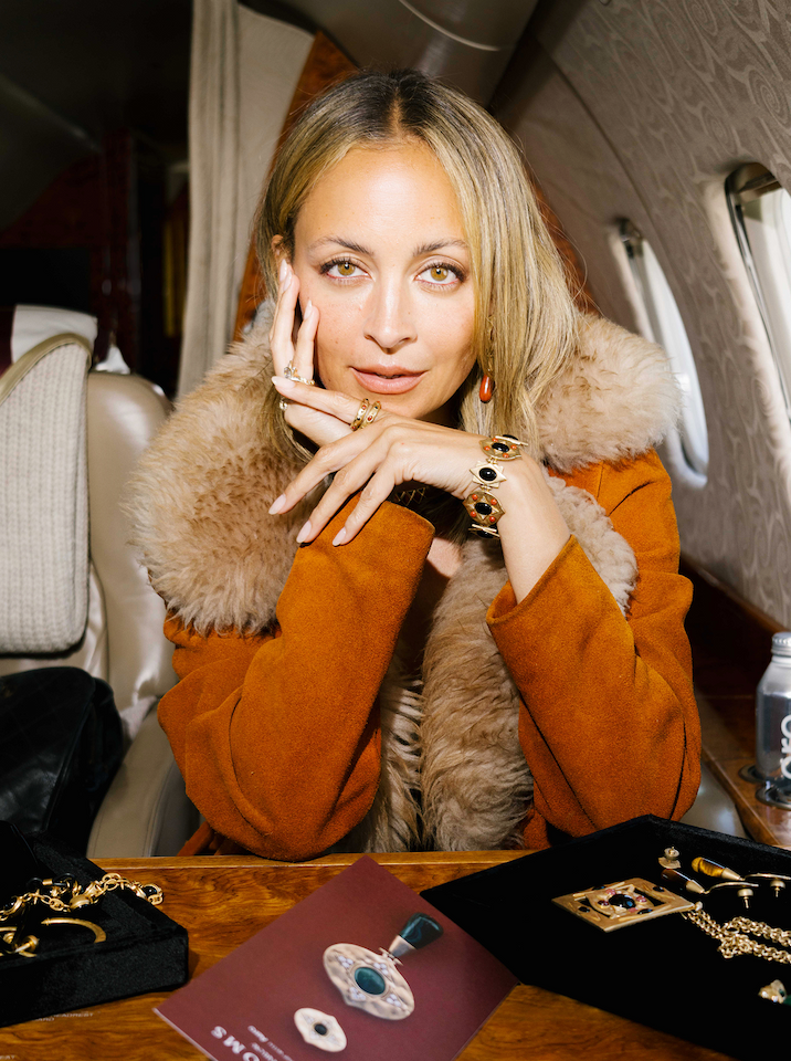 Nicole Richie dressed in a brown coat with a light brown fur trim, sits in a private Aero seat. Visible on her wrist is the Hathor Hinged Bracelet adorned with black and burgundy natural stones and set in plated gold. On her hands, gold rings. In front of Nicole are examples of House of Harlow jewelry for sale on the AIRLOOM flights.