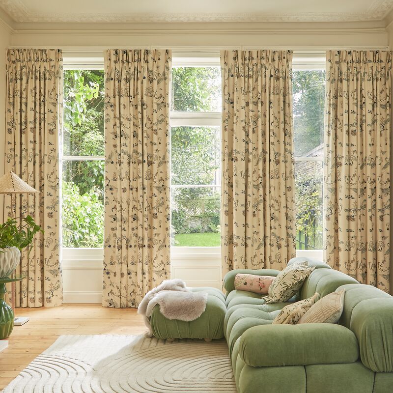 Floral curtains frame tall windows overlooking a leafy garden, bringing pattern and softness to a bright living room with a green sofa.