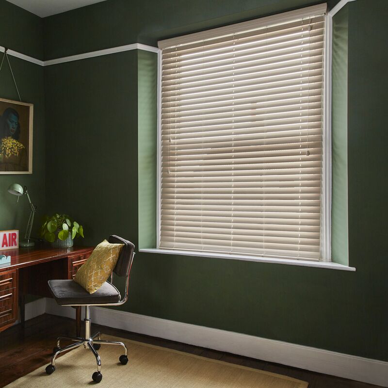 White Venetian blind fitted within a recessed window, bringing soft light to a dark green home office with a wooden desk and houseplants.