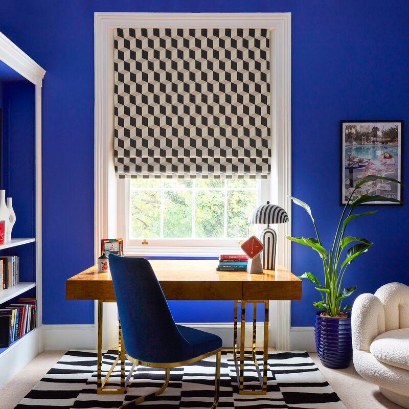 Geometric grey and cream Roman blind at a window, creating a bold focal point in a vibrant blue home office with a wooden desk.