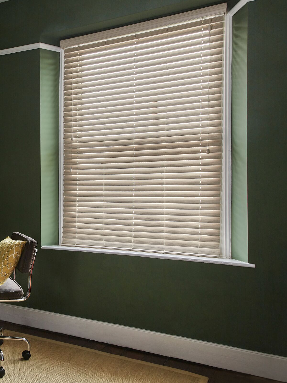White Venetian blind fitted within a recessed window, bringing soft light to a dark green home office with a wooden desk and houseplants.
