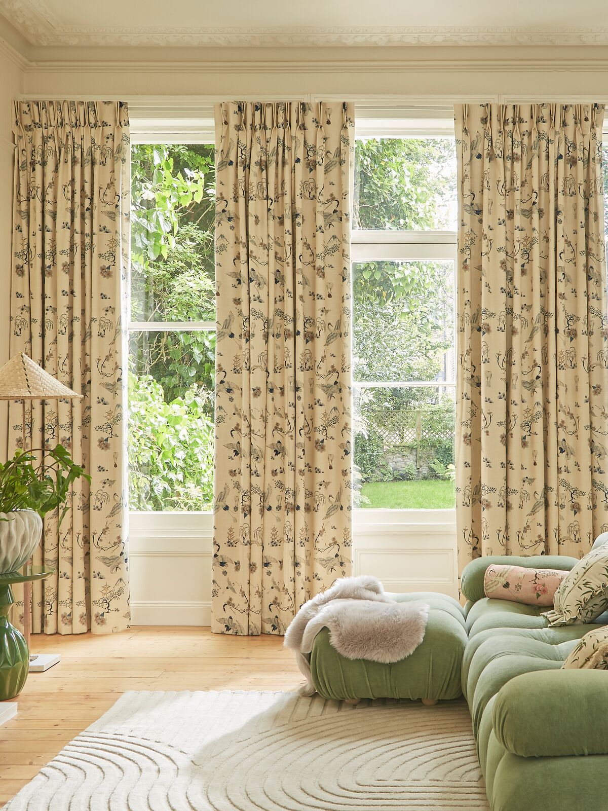 Floral curtains frame tall windows overlooking a leafy garden, bringing pattern and softness to a bright living room with a green sofa.