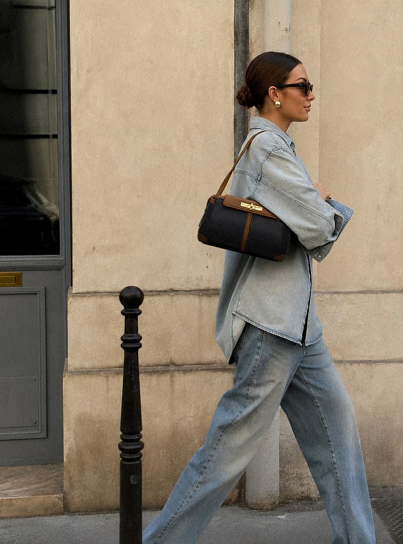 Model in a light-wash denim outfit carrying an East End Charlotte sml, black leather shoulder bag with rich brown trim and gold hardware.