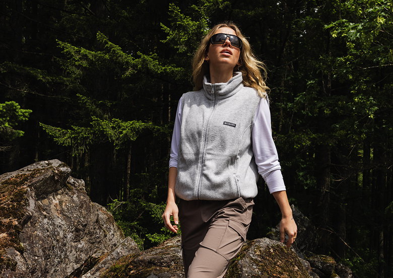 A woman hiking outside, wearing a white long sleeve shirt, vest, and pants.