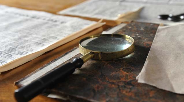 A magnifying glass placed on a textured book cover, surrounded by scattered sheets of paper on a wooden surface.