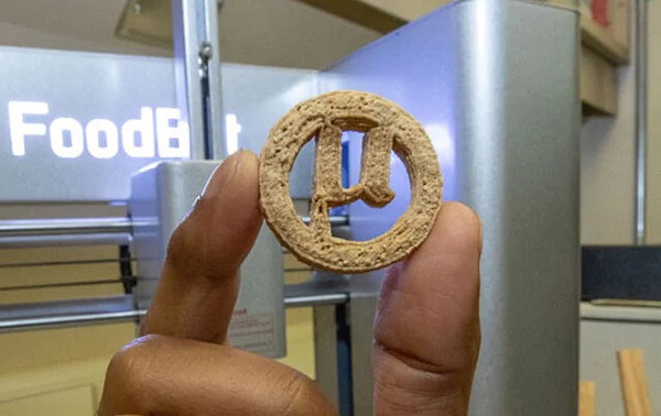 The 3D printed cookie being held between thumb and forefinger, with the logo visible.