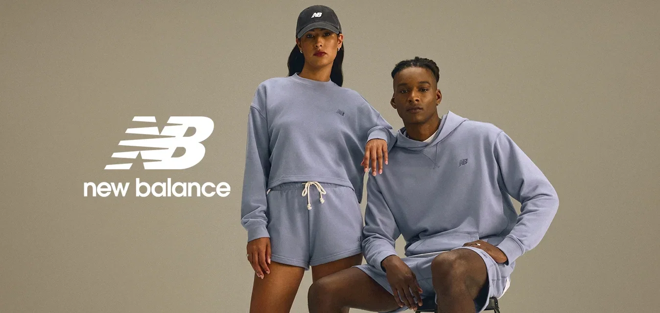 Twee modellen in bijpassende lichtpaarse sportkleding; de ene staat met een pet, de andere zit op een zwarte kruk, beide dragen witte sneakers.