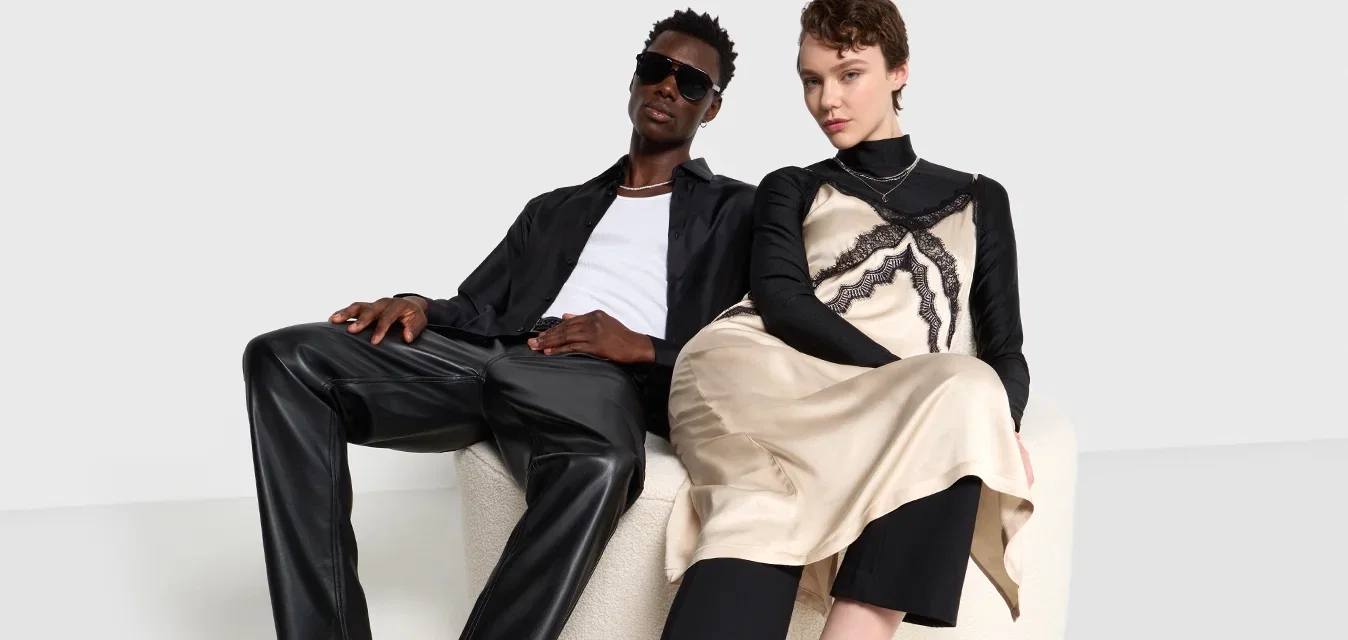 Twee modellen zitten op een ronde beige stoel. De één draagt een zwart topje met een patroon en een beige rok, de ander een zwart shirt, leren broek en schoenen.