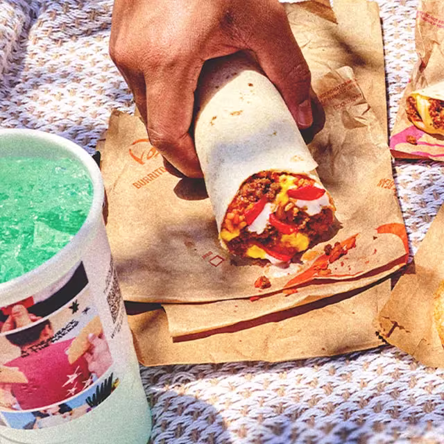 A person picking up a $2 Beefy Crunch Burrito from it's wrapper. There is a Medium Mountain Dew® Baja Blast™ on the table.