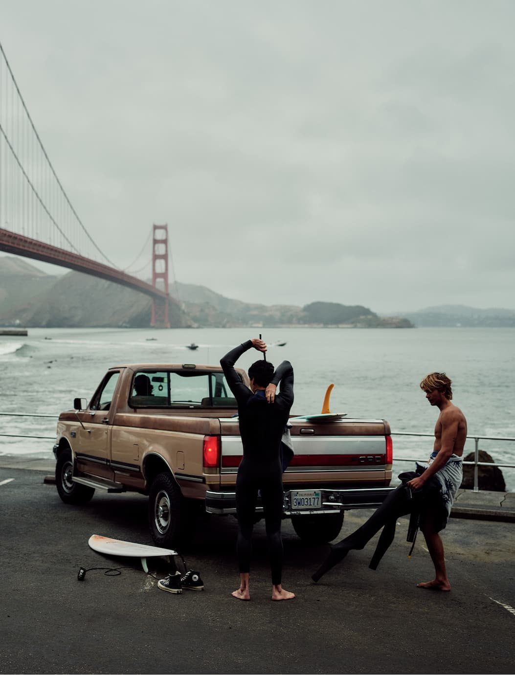 Two surfers gear up in wetsuits beside a truck with the Golden Gate Bridge in the background.