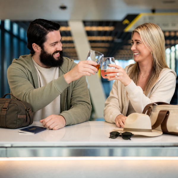 A man and woman sit at a bright airport bar, smiling as they clink glasses filled with drinks. Travel accessories including a passport, small bags, a hat, and sunglasses rest on the counter in front of them.