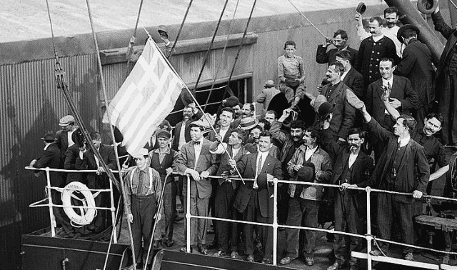 A group of men on a ship wave and hold a large flag, while others look on. The scene appears celebratory with a mix of formal and casual attire among the individuals.