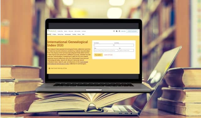 A laptop displaying the International Genealogical Index website sits on an open book, with stacks of books and blurred library shelves in the background.
