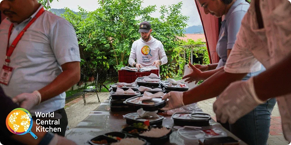 Image Description: World Central Kitchen crew members packing and preparing to-go meals at a long table with greenery in the background. World Central Kitchen logo in the bottom left corner of the image.