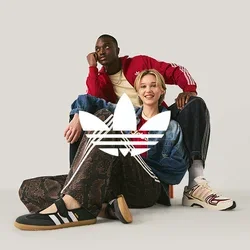 Een man in een rode jas en een vrouw in een spijkerjasje en Adidas-broek zitten op de vloer tegen een effen lichte achtergrond.