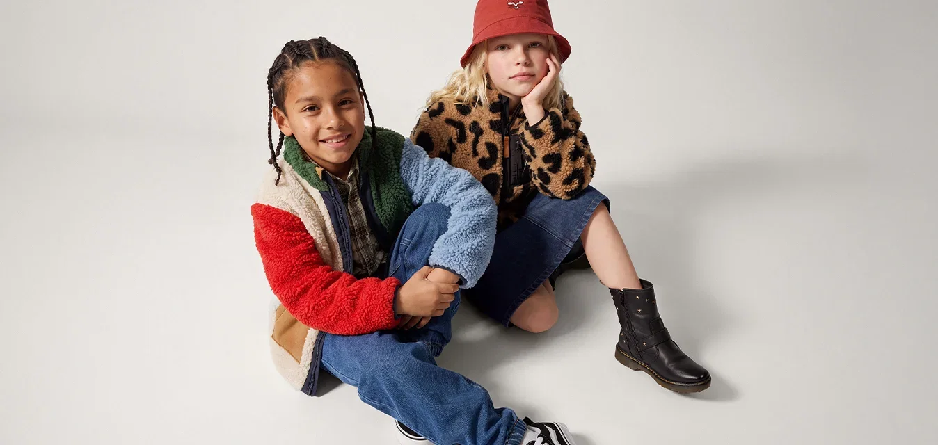 Twee kinderen zitten op de grond, de een in een multicolor fleecejack en de ander in een jas met luipaardprint en een rode emmerhoed.